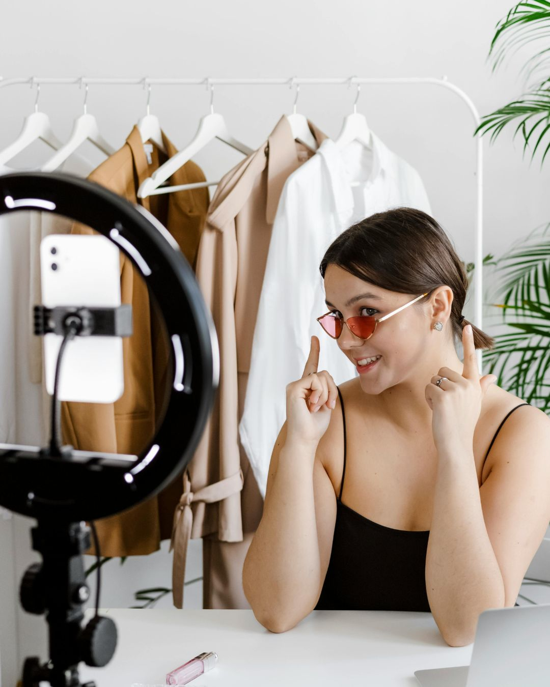 Small business owner creating a social media video with a smartphone and ring light, promoting fashion products in front of a clothing rack.