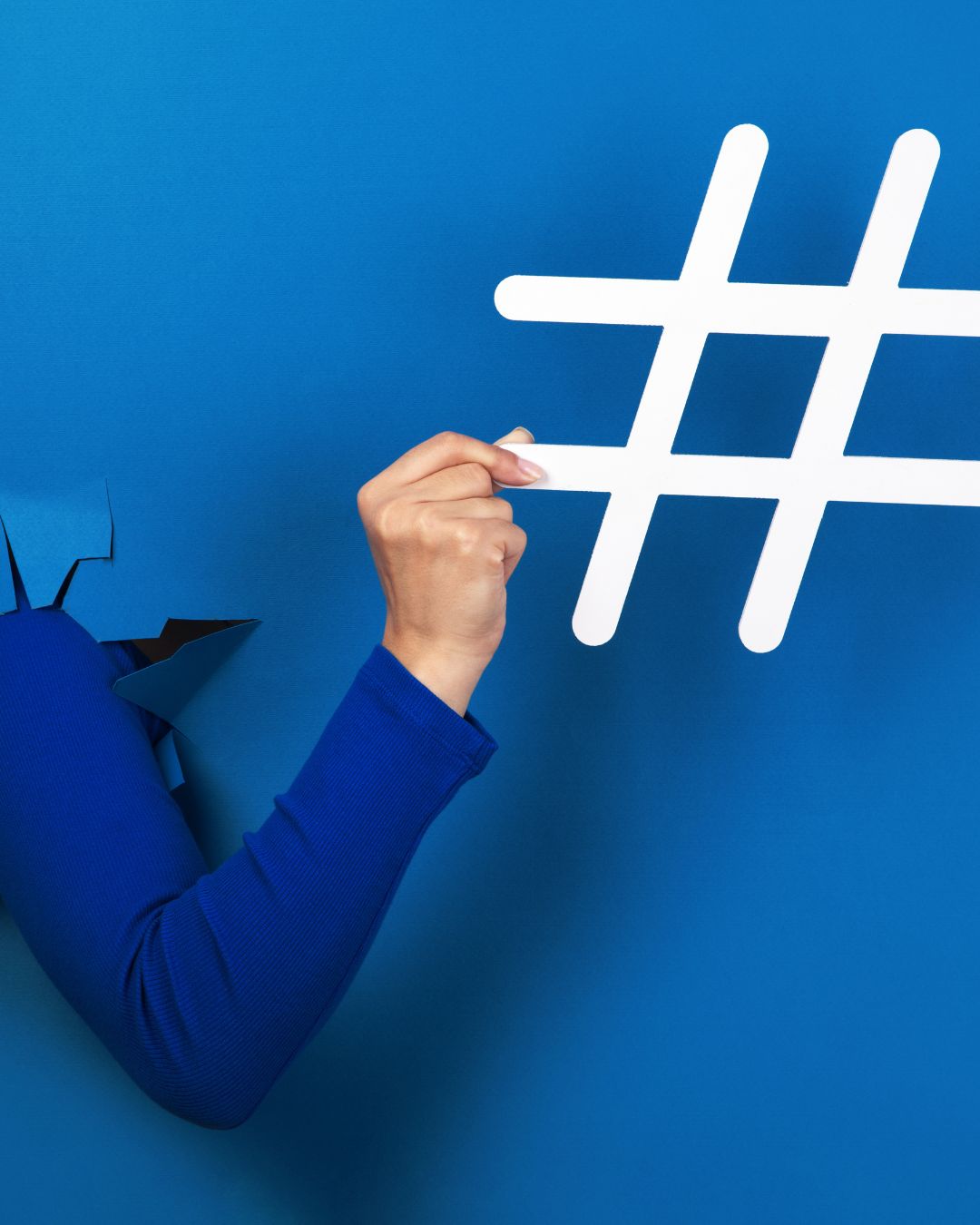 Hand in blue sleeve breaking through a backdrop while holding a large white hashtag symbol — highlighting the power of hashtags for local businesses.