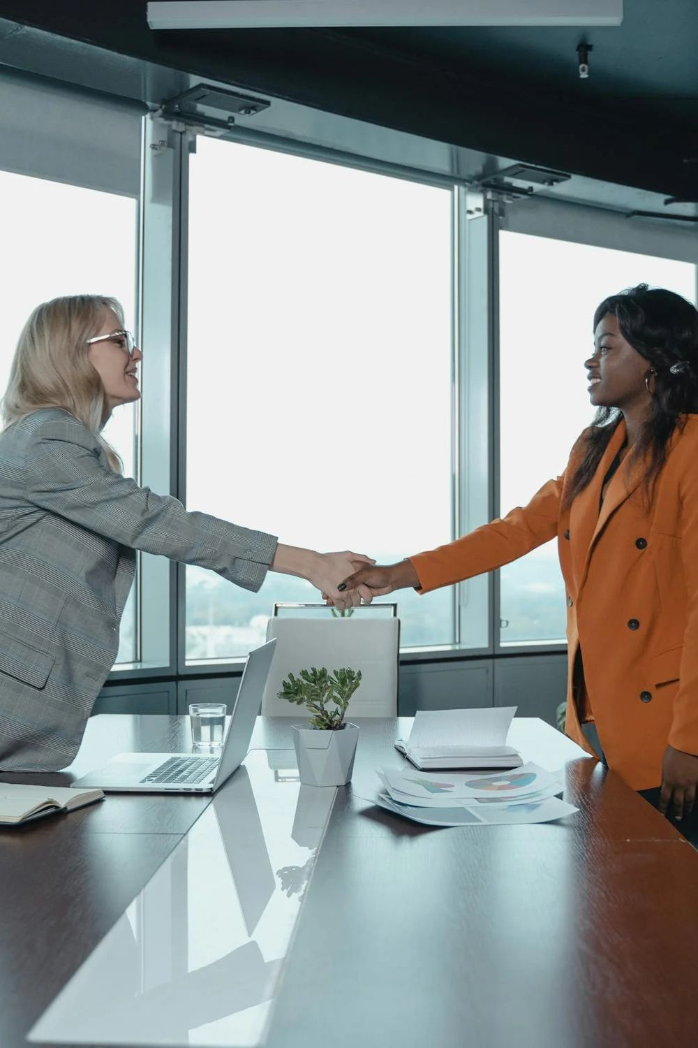 Two businesswomen shaking hands across a conference table in a modern office
