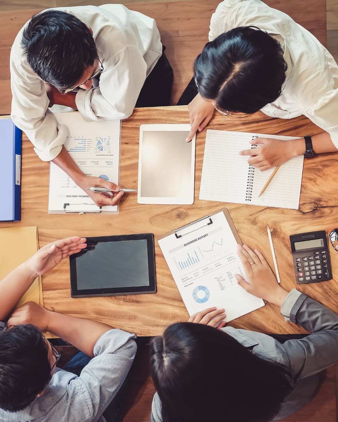 Overhead view of four colleagues seated around a wooden table with two tablets, printed business reports displaying charts and graphs, an open notebook, and a calculator as they collaborate on social media and website performance.