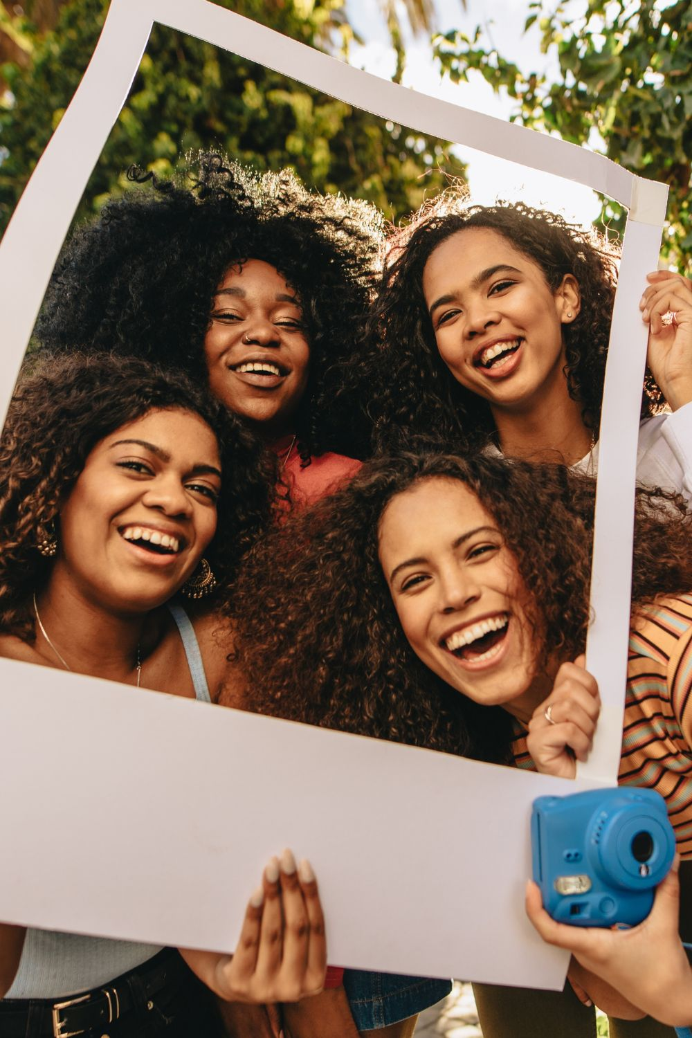 Four friends smiling and posing together outdoors while holding a photo frame prop.