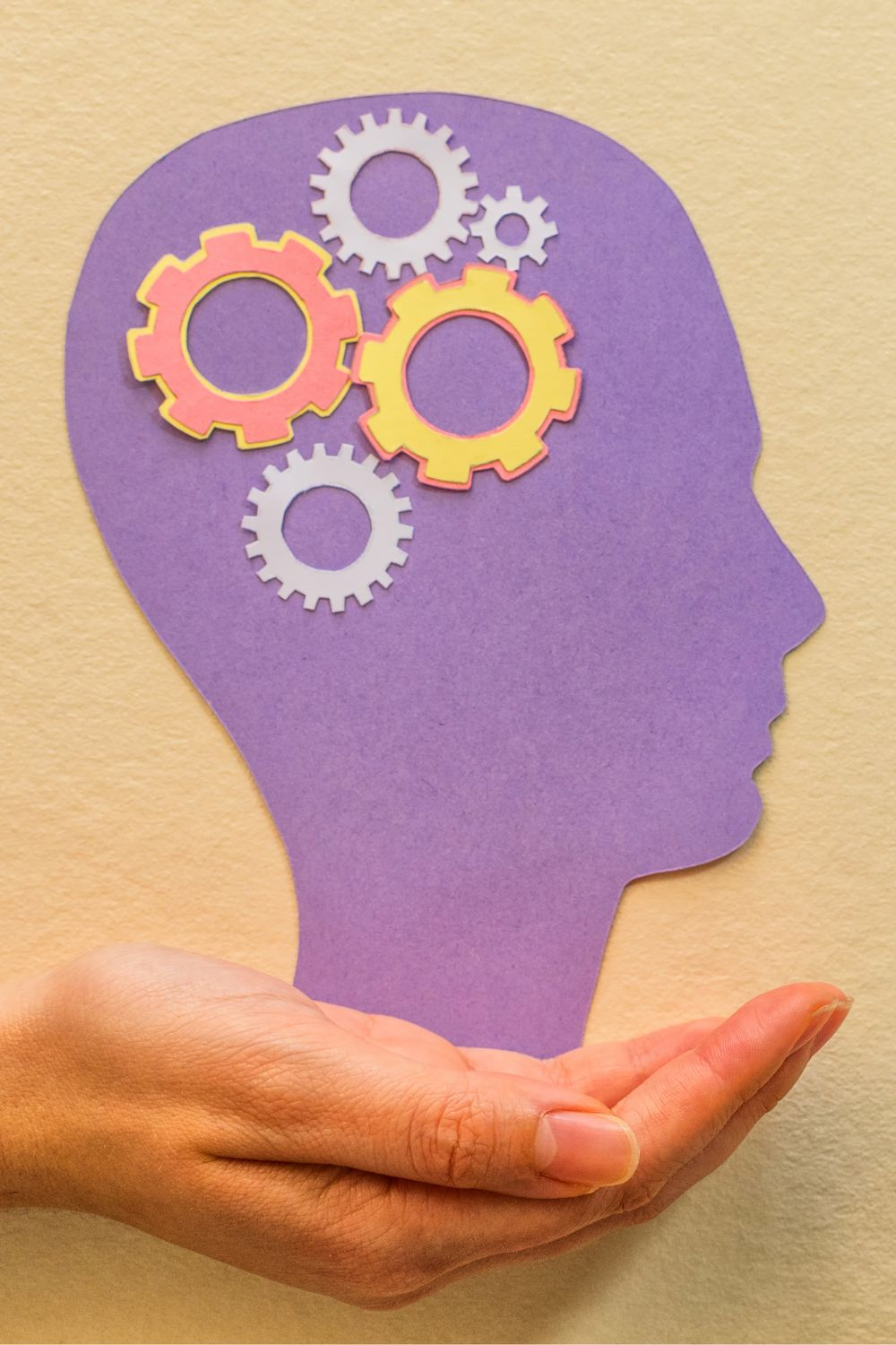 Purple paper cutout of a human head with colorful gear icons inside, symbolizing thinking and strategy, held by a hand.