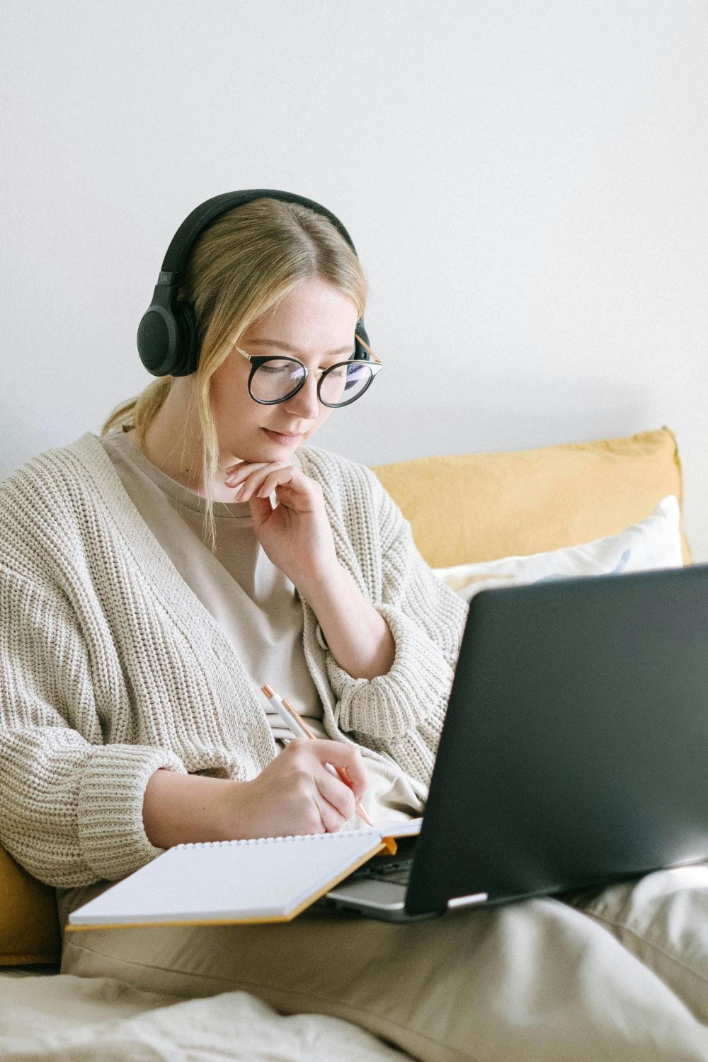 Woman wearing headphones while working on a laptop and writing notes in a notebook from home.