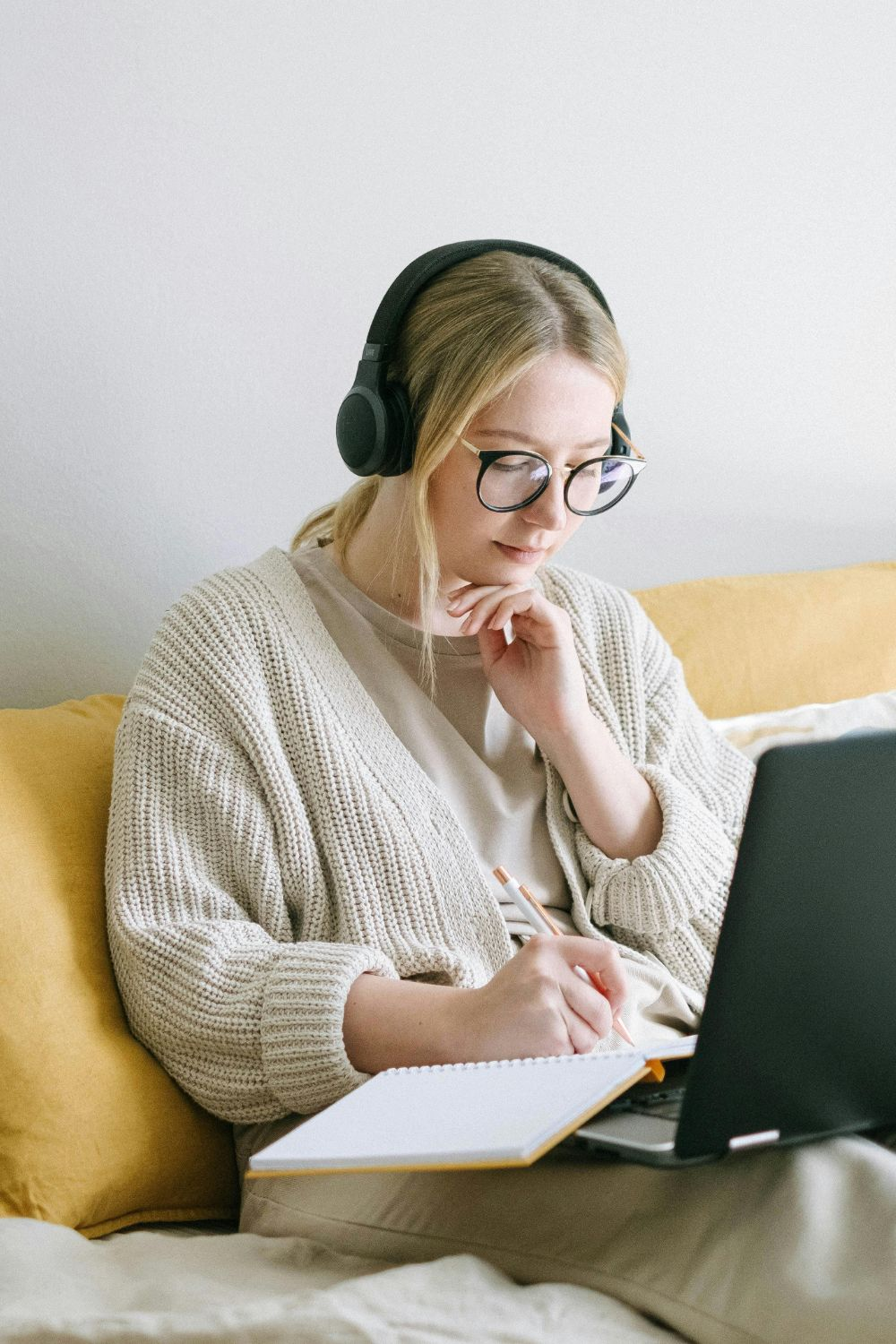 Woman wearing headphones and glasses taking notes while working on a laptop at home.