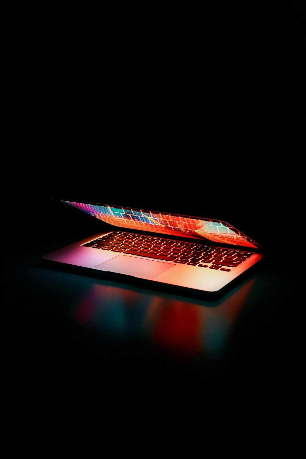 Half-open laptop glowing with colorful neon light in a dark room.