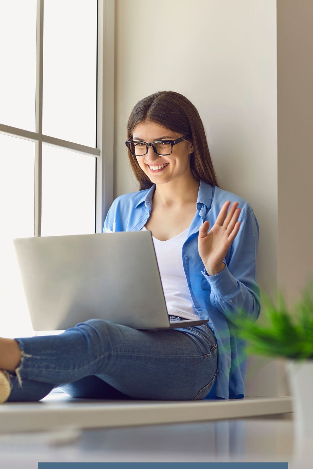 Young woman waving at her laptop during a virtual meeting, sitting by a window and smiling.