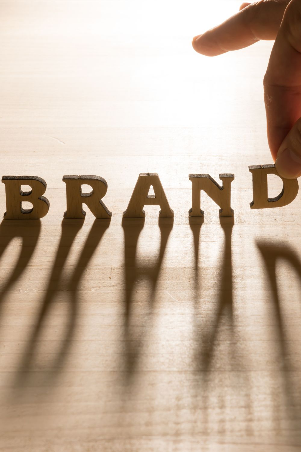 Hand arranging wooden letters spelling "BRAND" with long shadows on a wooden table, symbolizing branding strategy.