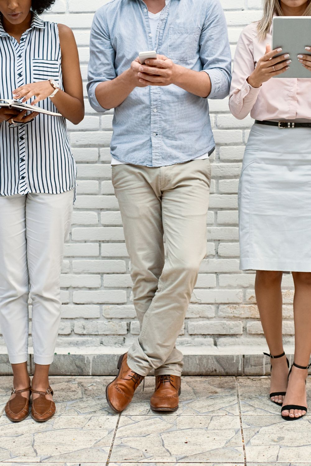 Three people standing against a white brick wall while using mobile devices and tablets